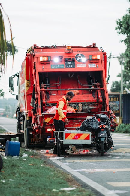 A sanitation worker wearing a high-visibility yellow and red uniform is operating a large red rubbish collection vehicle on a street during daytime. The worker is seen standing at the back of the vehicle, which has its rear compartment open, revealing a mechanical compactor and hydraulic system. The worker appears to be loading or managing waste, with a black garbage bag placed nearby on a small platform attached to the vehicle. The scene includes a street with a sidewalk and a grassy area, with some litter scattered on the ground. In the background, there are trees, power lines, and a few commercial signs, indicating an urban or suburban environment. The lighting is natural, suggesting an overcast day, and the wider setting emphasizes the routine of independent waste collection and rubbish removal services in Finchley, managed by Rubbish Removal Finchley.