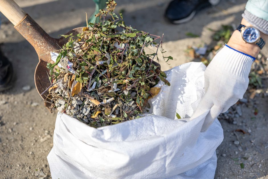 Several black rubbish bags filled with waste are scattered across a grassy area in front of a multi-storey residential building. The bags are made of thick plastic with a matte finish, and some are tightly sealed while others appear loosely tied. Behind the bags, a row of leafless trees with dark, textured bark partially obscures the building's facade, which features large windows and a combination of brickwork and paneling in shades of gray and white. The scene suggests an outdoor waste collection point, possibly related to private rubbish removal services, with the bags ready for disposal or collection. The environment is overcast, with diffuse natural light illuminating the scene evenly, creating a subdued and neutral tone. The overall composition indicates an area prepared for rubbish removal, consistent with service providers like Rubbish Removal Finchley handling waste from residential properties in the vicinity.