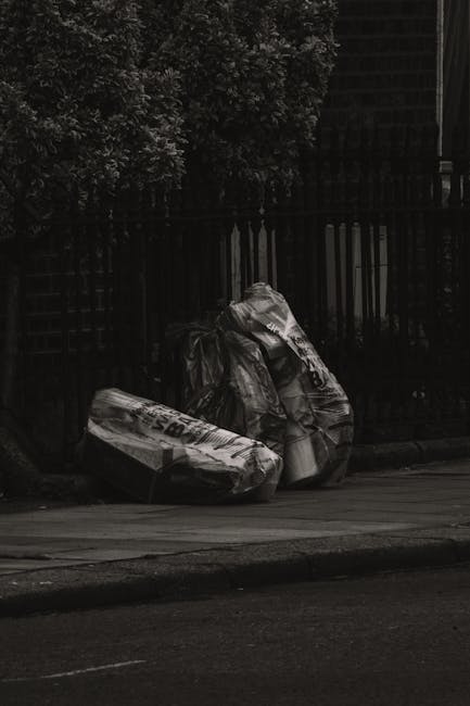 Three black rubbish bags filled with waste resting on the edge of a pavement in a quiet outdoor setting. The bags are made of thick plastic with a slightly crinkled texture and are tied at the top, showing some wrinkles and creases. They are positioned near a wooden fence with vertical slats, which is dark in colour, and behind the fence, there is dense foliage from trees or bushes, adding a natural background to the scene. The pavement appears smooth with a light colour, contrasting with the darker curb and road surface in the foreground. The overall atmosphere is subdued, possibly during overcast weather, with diffused lighting creating minimal shadows. This scene suggests an instance of private rubbish disposal or collection, relevant to waste management services offered by companies like Rubbish Removal Finchley, especially in contexts requiring alternative or independent waste handling outside local authority collection processes.