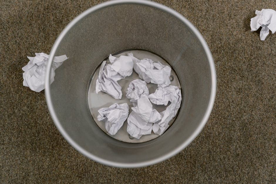 An overhead view of a cylindrical metal rubbish bin placed on a brown textured carpeted floor. Inside the bin, there are several crumpled white paper sheets submerged in a small pool of water. Additional crumpled paper pieces are scattered on the carpet around the bin, indicating recent waste disposal. The metal surface of the bin has a matte finish, with a smooth, slightly reflective interior. The scene depicts a clean, indoor environment with natural lighting highlighting both the metallic sheen of the bin and the crumpled paper textures. The image is relevant to independent rubbish collection or onsite waste handling, as it visually emphasizes waste disposal practices suitable for commercial settings such as shops or offices, aligning with the services provided by Rubbish Removal Finchley.