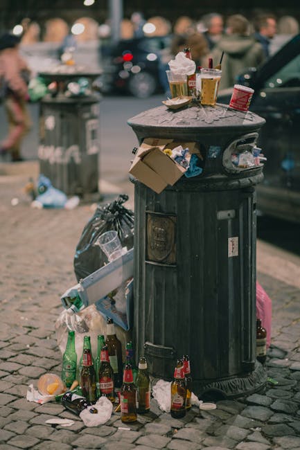 A black metal public rubbish bin situated on a cobblestone street with its lid closed, surrounded by overflowing litter and discarded bottles, including glass beer bottles and crumpled paper, scattered at its base. On top of the bin, there are disposable coffee cups with plastic lids and a red takeaway coffee cup, along with a small cardboard box, indicating improper disposal of food and beverage waste. The environment appears to be an urban outdoor area, with a blurred background showing parked cars and a busy street scene, including pedestrians and additional rubbish bins. The scene highlights issues related to waste management and the need for effective rubbish removal services, such as those provided by Rubbish Removal Finchley, to manage street cleanliness and curb littering. Lighting conditions suggest natural daylight, casting soft shadows on the cobblestones, emphasizing the disorganized state of the refuse around the bin.