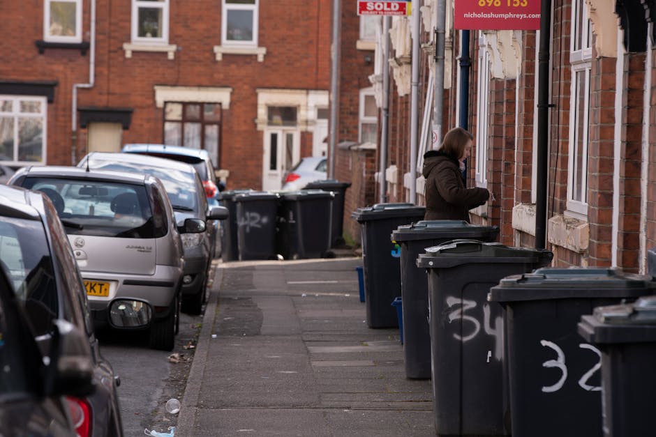 A residential street scene featuring a row of dark grey wheelie bins with white spray-painted numbers placed along the concrete sidewalk in front of brick terraced houses. The bins are positioned close to the building facades, which have white-framed windows and repair patches on the brickwork. In the background, a woman dressed in a dark brown coat is seen with her head bent over, possibly interacting with one of the bins or the house entrance. Several parked cars of various makes and colours occupy the street, extending from the foreground to the background, with some cars partially obscured by the bins. The environment is typical of an urban area with a mix of residential and waste disposal activity, illustrating an on-site collection or private rubbish handling scenario, with the scene well-lit by natural daylight. Rubbish Removal Finchley services this general type of waste management, handling waste within tight access properties, as evidenced by the closely positioned bins and narrow street setting.