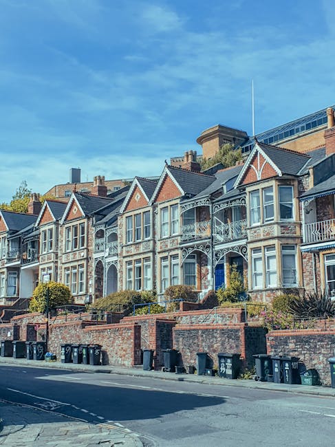 A row of Victorian-style residential buildings with ornate cast-iron balconies and large bay windows, set on a slope with brick retaining walls and small landscaped front gardens. The facades feature a mix of red brick and decorative white trim, with steep gable roofs covered in dark slate tiles. In the foreground, a paved street with black wheelie bins lined up along the curb indicates waste disposal. The base of the buildings is bordered by low brick walls, some topped with concrete coping stones, separating the front gardens from the pavement. The scene is well-lit by natural daylight, with a clear blue sky and a few wispy clouds overhead, suggesting a calm, sunny day. The overall setting depicts an urban residential area where waste collection might be handled either through local authority services or private providers such as Rubbish Removal Finchley, especially for properties with limited access such as those on this terraced street.