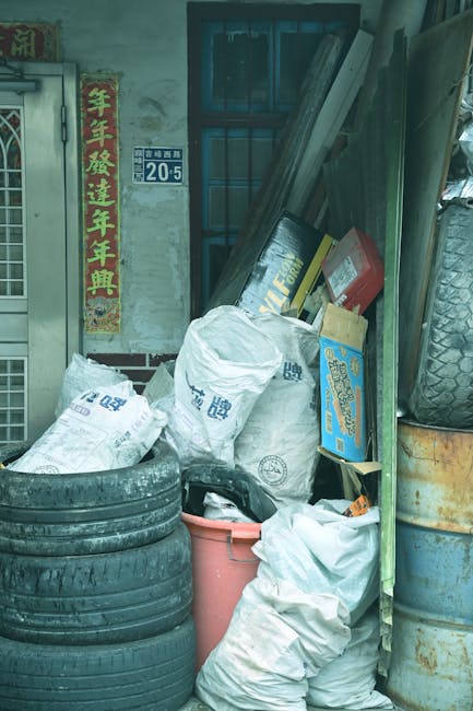 A collection of used car tires stacked in a corner outside a building, with several large white plastic bags filled with rubbish placed nearby. A red plastic bucket and a smaller black bag are also visible in front of the bags. Leaning against the building's wall are long wooden planks and cardboard boxes, some labelled with text and logos. The background shows a window with metal bars and a partially visible door with a metallic frame, alongside a brick wall. To the right, an upright tyre is positioned beside a weathered, rusty metal cylinder. The environment appears cluttered and untidy, representing typical waste or junk accumulation that a rubbish removal service such as Rubbish Removal Finchley might collect during an on-site clearance. The scene is lit by natural ambient light, showing signs of outdoor storage, and the setting highlights the need for professional rubbish removal or private disposal services in an urban area in Finchley.