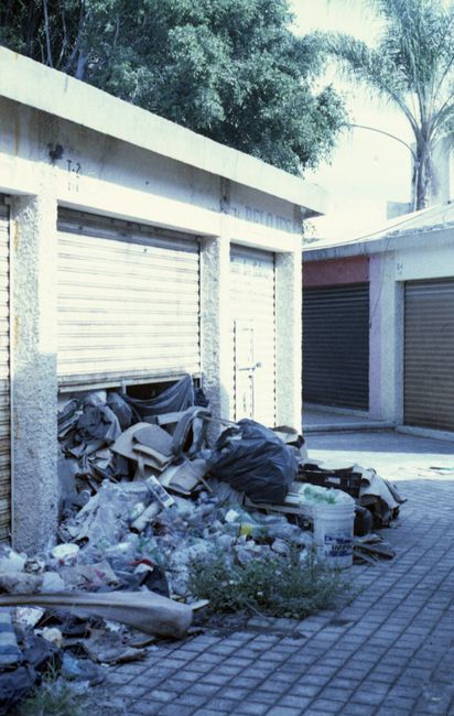 An outdoor scene showing a small, concrete garage area with three roller shutter doors; two are closed, painted silver, and one is partially open, revealing dark interior. In front of the open garage, there is a significant accumulation of mixed waste materials including black rubbish bags, broken and discarded cardboard boxes, plastic packaging, and various debris. The waste appears to be a combination of household and building rubbish, strewn on a paved surface composed of small, rectangular bricks. In the background, tall trees with dense foliage and slim tree trunks are visible, casting shadows over the area. The environment suggests an untidy, neglected space that could benefit from professional rubbish removal services, such as those provided by Rubbish Removal Finchley, to facilitate on-site clearance and proper disposal of the material.