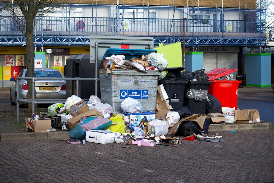 A collection of used car tires stacked in a corner outside a building, with several large white plastic bags filled with rubbish placed nearby. A red plastic bucket and a smaller black bag are also visible in front of the bags. Leaning against the building's wall are long wooden planks and cardboard boxes, some labelled with text and logos. The background shows a window with metal bars and a partially visible door with a metallic frame, alongside a brick wall. To the right, an upright tyre is positioned beside a weathered, rusty metal cylinder. The environment appears cluttered and untidy, representing typical waste or junk accumulation that a rubbish removal service such as Rubbish Removal Finchley might collect during an on-site clearance. The scene is lit by natural ambient light, showing signs of outdoor storage, and the setting highlights the need for professional rubbish removal or private disposal services in an urban area in Finchley.